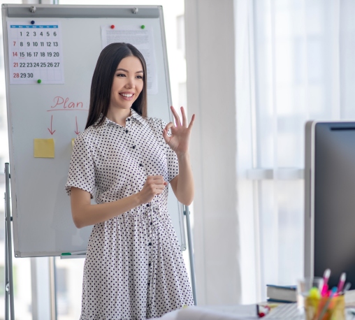 A woman confidently displays the "OK" hand gesture while standing in front of a whiteboard.