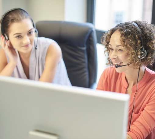 Two professional women wearing headsets, focused on their computer screen as they collaborate and work together.